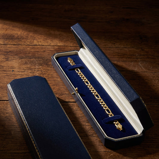 A gold-toned chain bracelet displayed inside an open dark blue jewelry box with a white interior, placed on a wooden surface. The matching closed box is visible in the foreground.